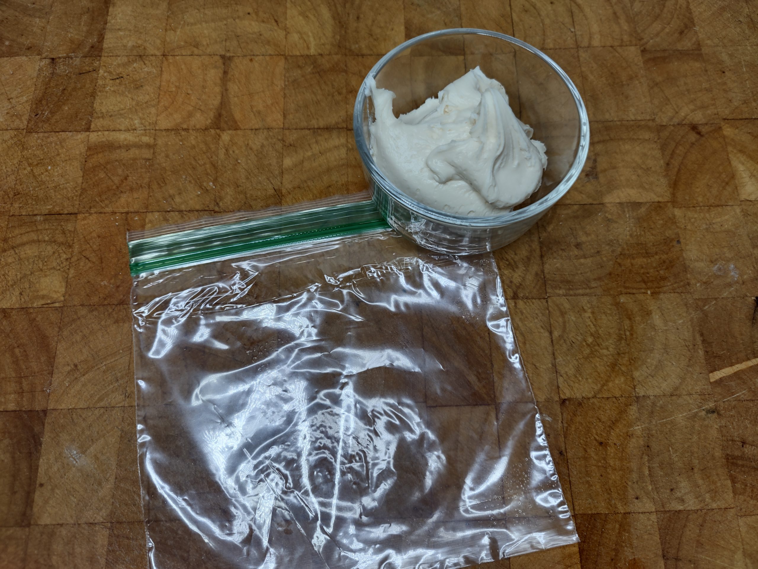 bowl of cream cheese frosting sitting next to freezer bag on a wooden table