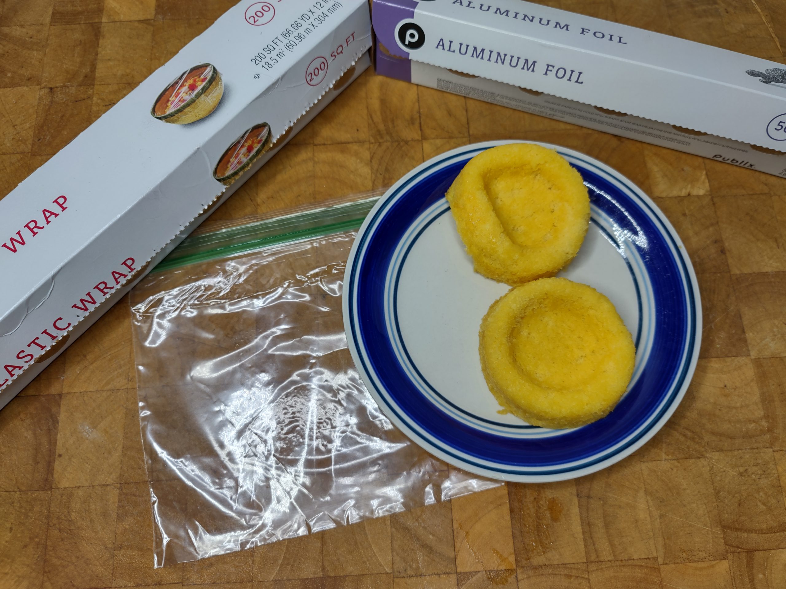two slices of sponge cake on a plate next to an empty freezer bag, roll of plastic wrap and roll of aluminum foil