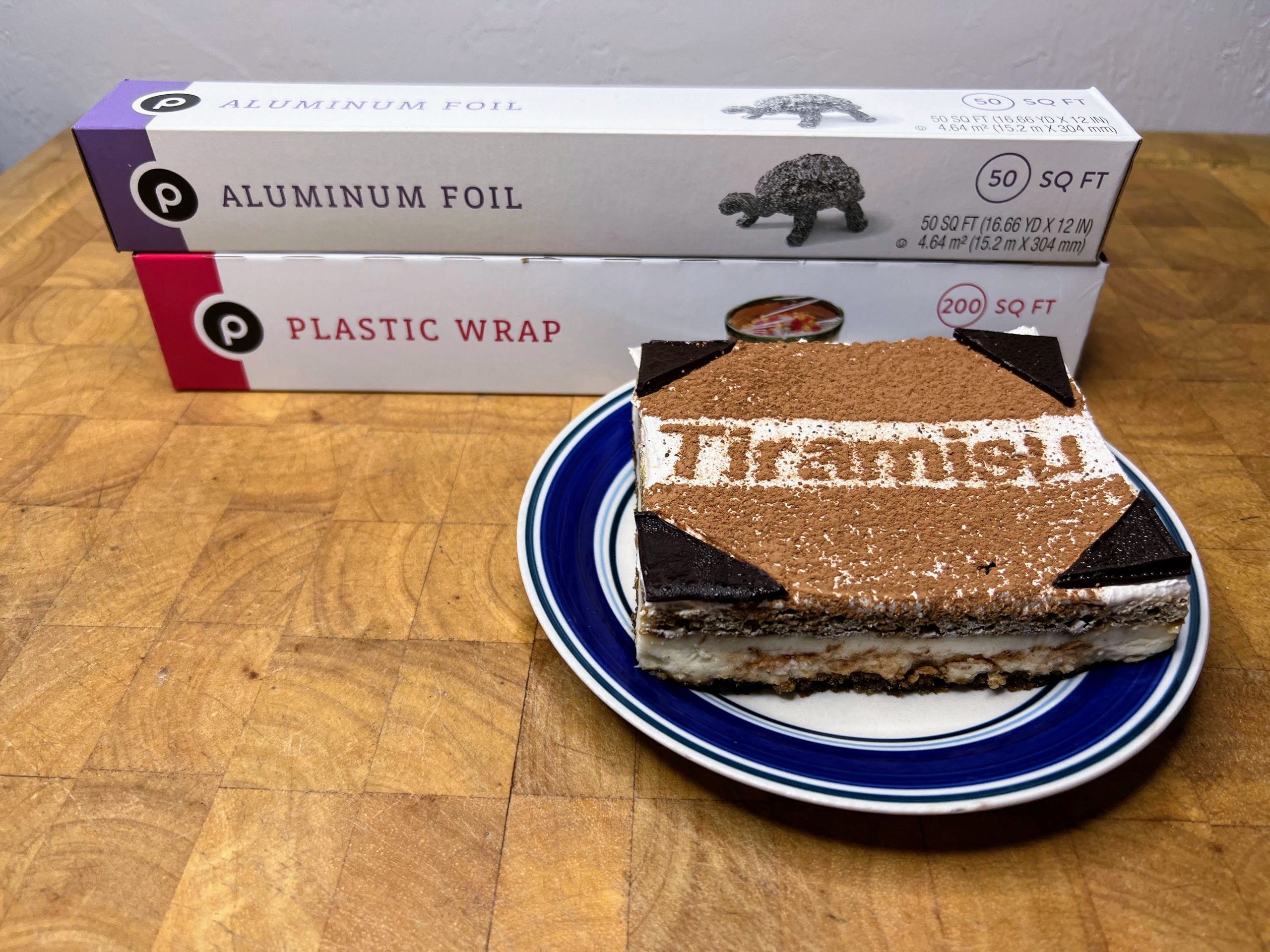 slice of tiramisu on blue and white plate with plastic wrap and aluminum foil next to it