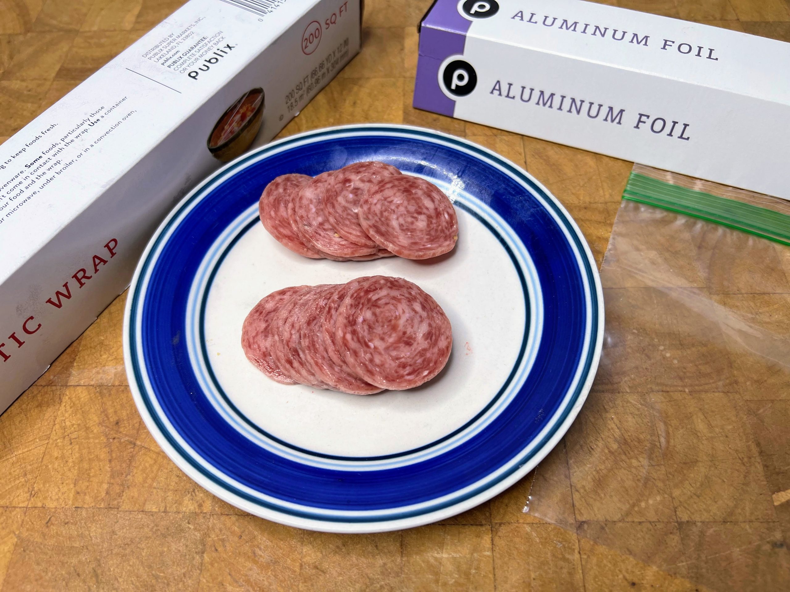 stacks of salami on a plate next to empty freezer bag, aluminum foil and plastic wrap
