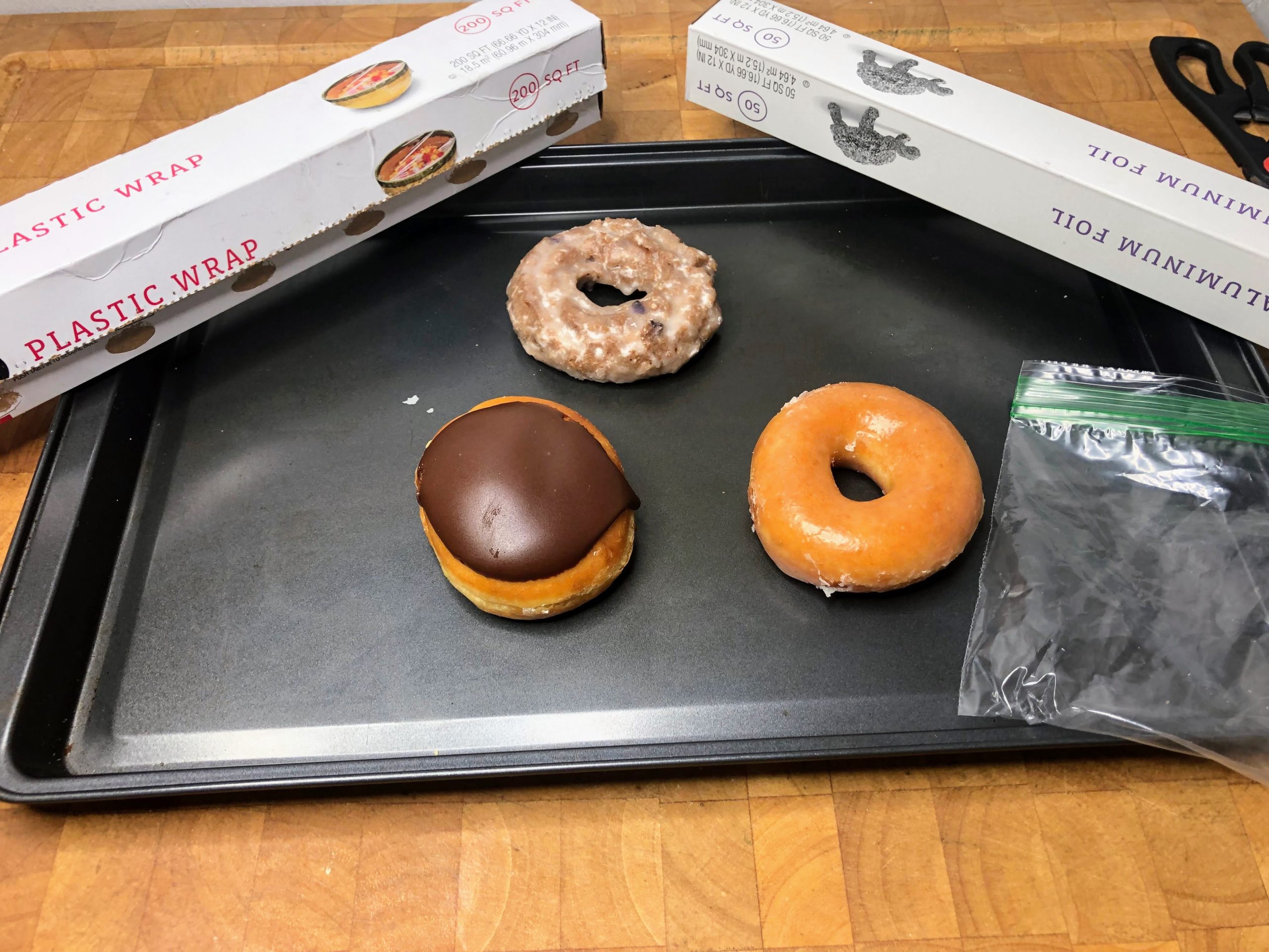 three krispy kreme doughnuts on a cookie sheet with an empty freezer bag, aluminum foil and plastic wrap