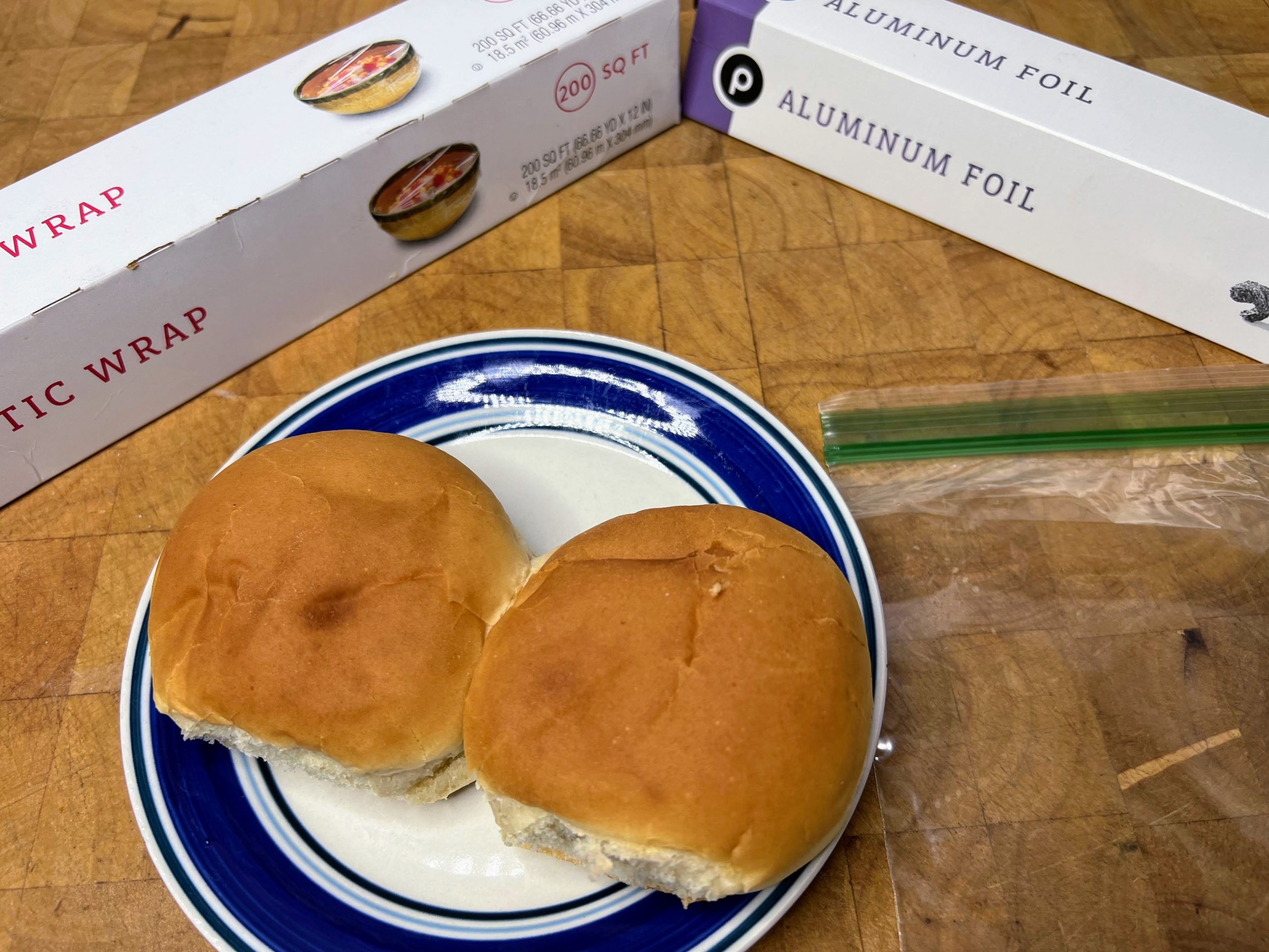 two hamburger buns on a plate next to an empty freezer bag, plastic wrap and aluminum foil