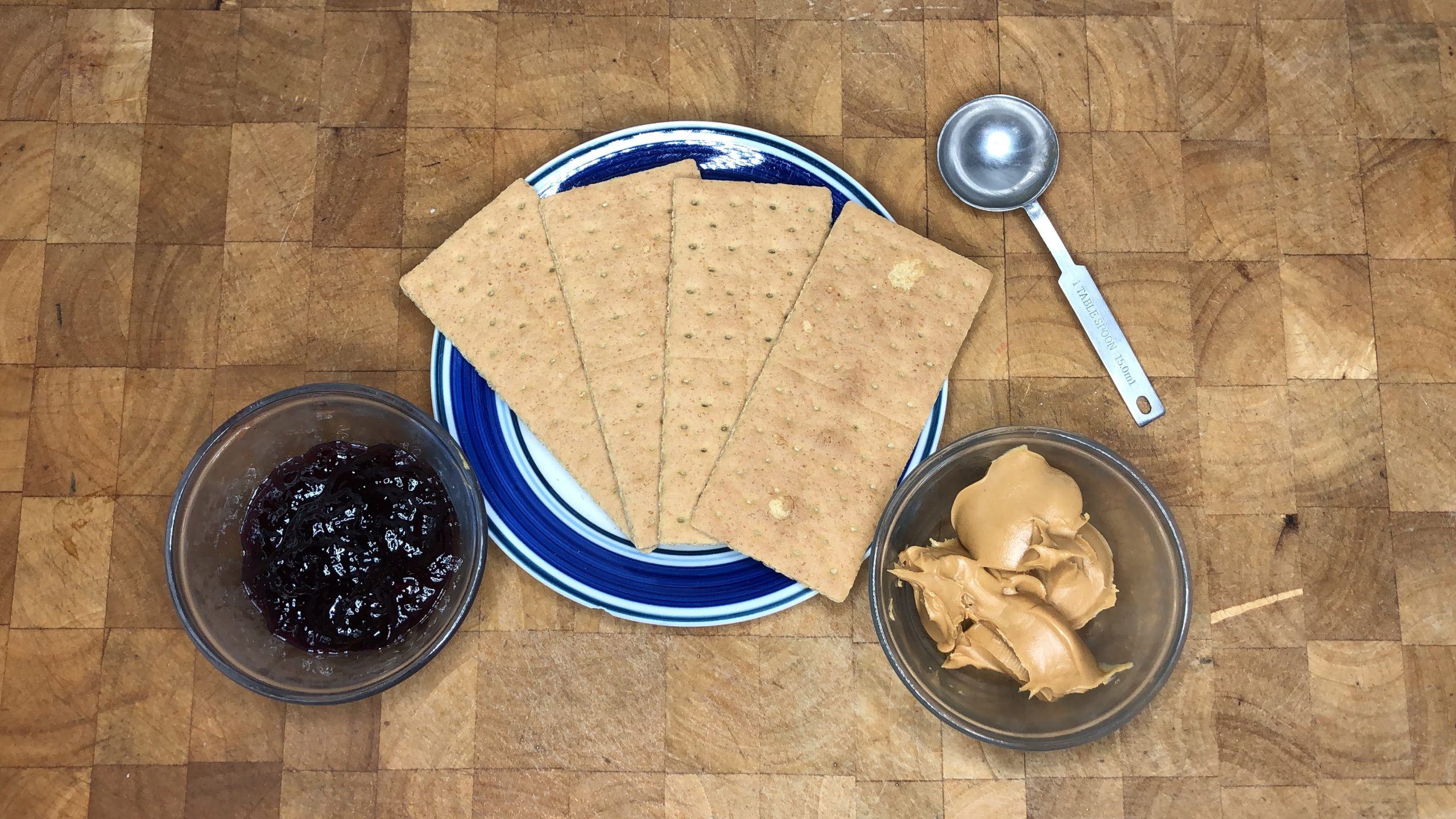 Four graham crackers on a plate next to a measuring spoon and bowls of peanut butter and jelly.